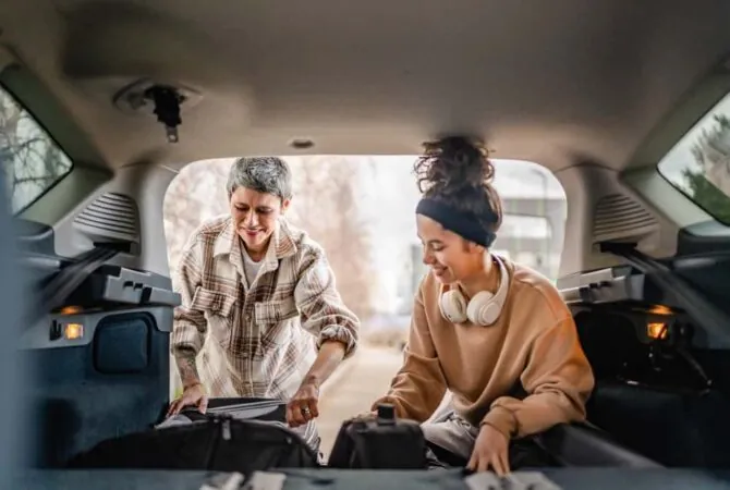 A parent and teenager pack the trunk of a car with items for college. The photo was taken from inside the car to show the parent and teenager outside, loading items into the trunk.