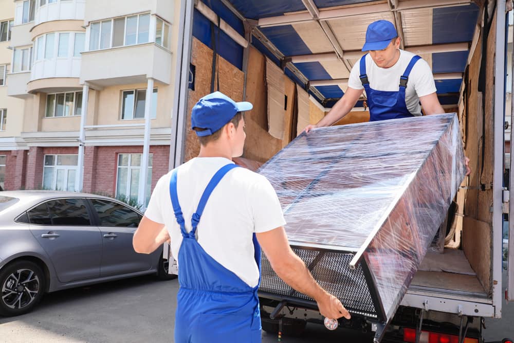 Two professional movers in blue coveralls and hats loading metal rack into empty moving truck outside townhome