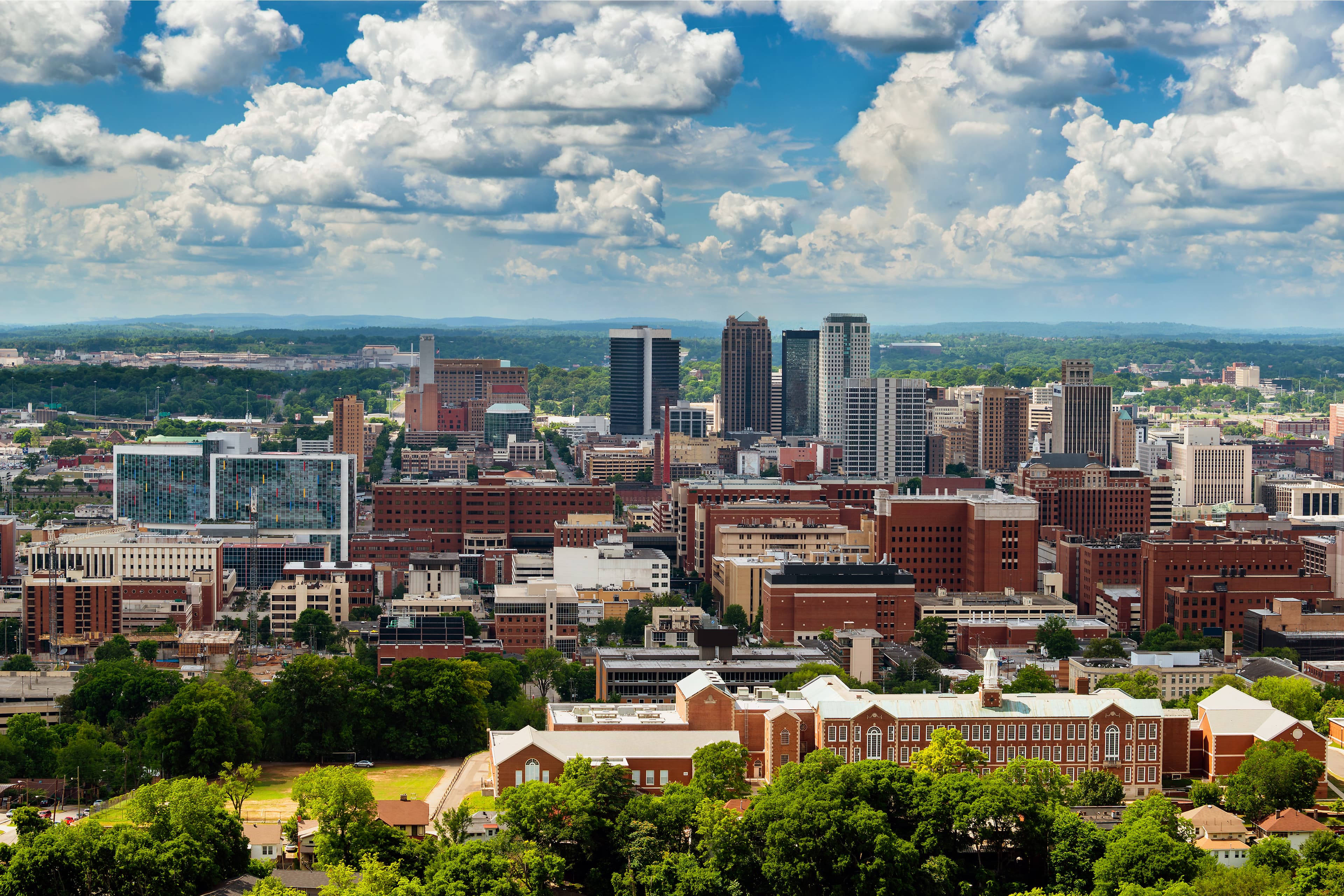 Photo of the Birmingham, AL skyline, featuring tall buildings and green trees during the day.