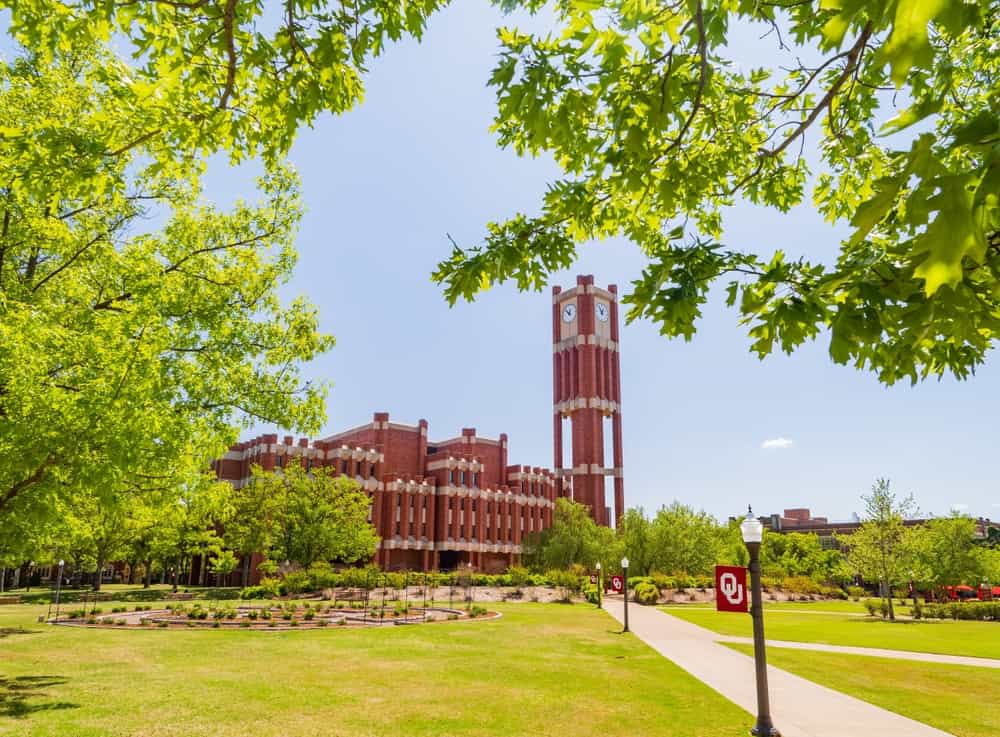 View of the University of Oklahoma campus in Norman, featuring red-brick academic buildings and a clock tower surrounded by a green lawn and trees.