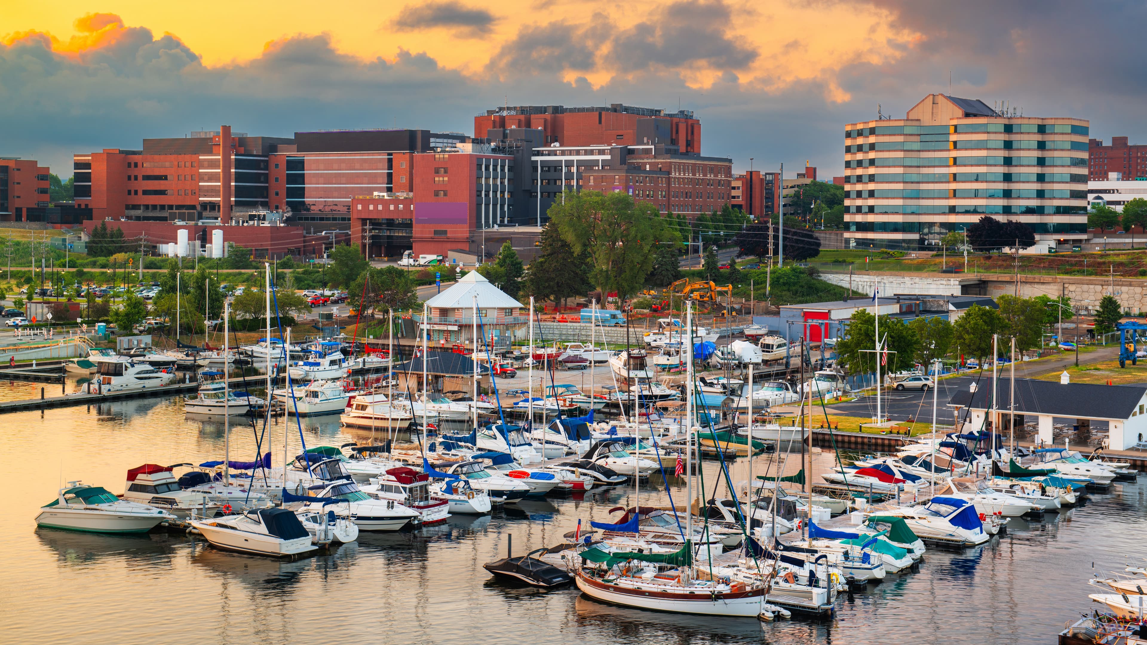 A sunset photo of Erie, PA with large brick buildings in the background, greenspace, and boats at docks.