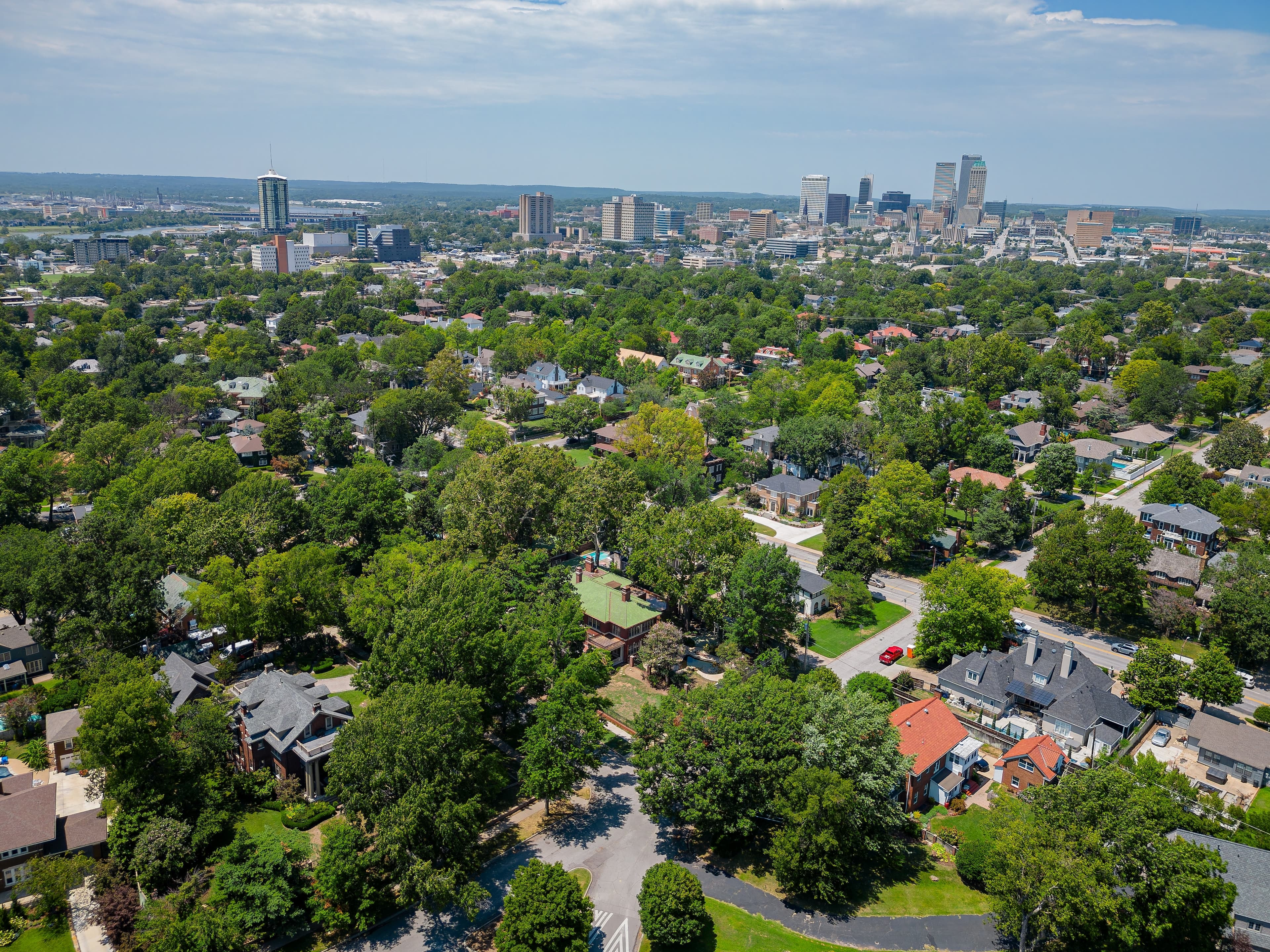 Overhead photo of Broken Arrow featuring colorful houses on tree-lined streets with the Tulsa skyline in the background on a sunny day.