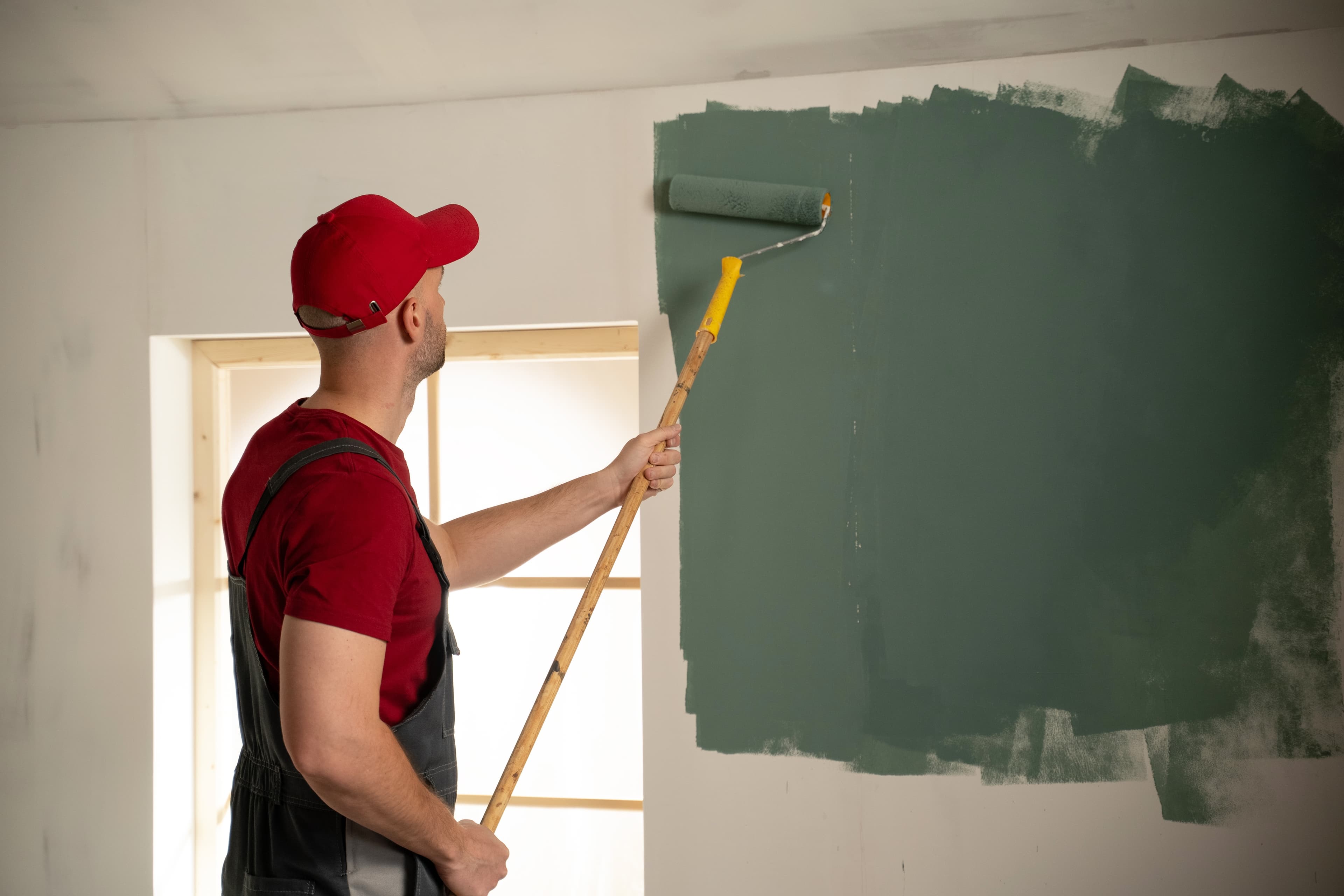 Painter using a roller on an extension pole to apply green paint to an interior wall