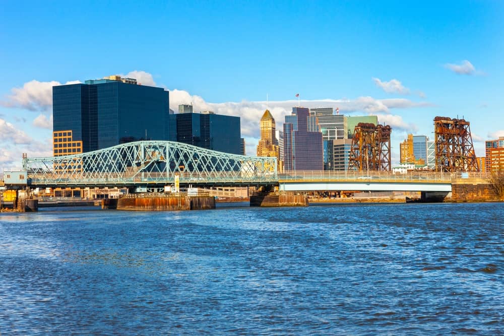 Skyline shot of Newark, NJ along a riverfront during day-time, featuring blue skies and fluffy clouds.