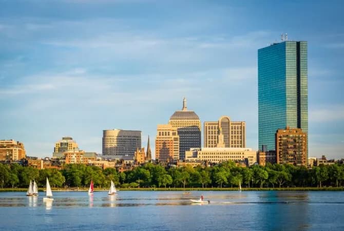Boston skyline on clear day with water and sailboats in foreground and buildings and trees in background
