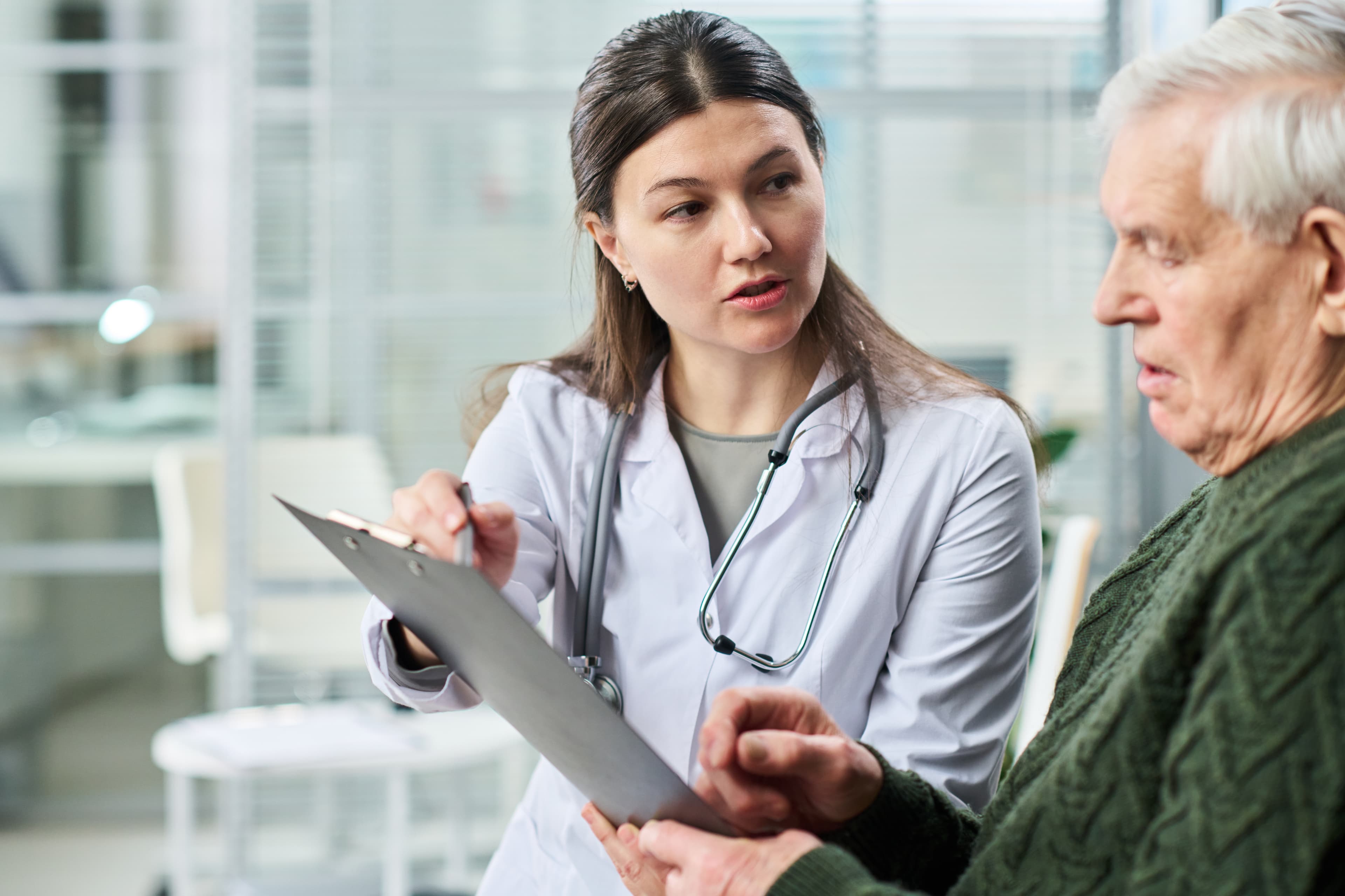 Doctor in a white coat pointing to a clipboard while discussing records with an older patient