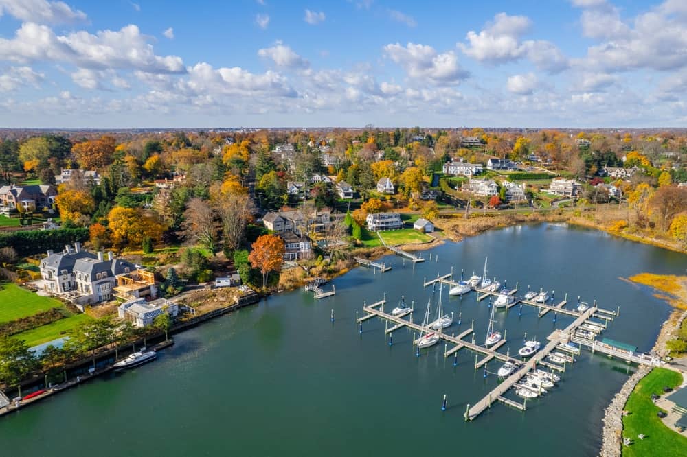Aerial photo of Stamford, CT during daytime, featuring blue skies and fluffy clouds. Also featured are autumn leaves and a large body of water with boats and a marina.