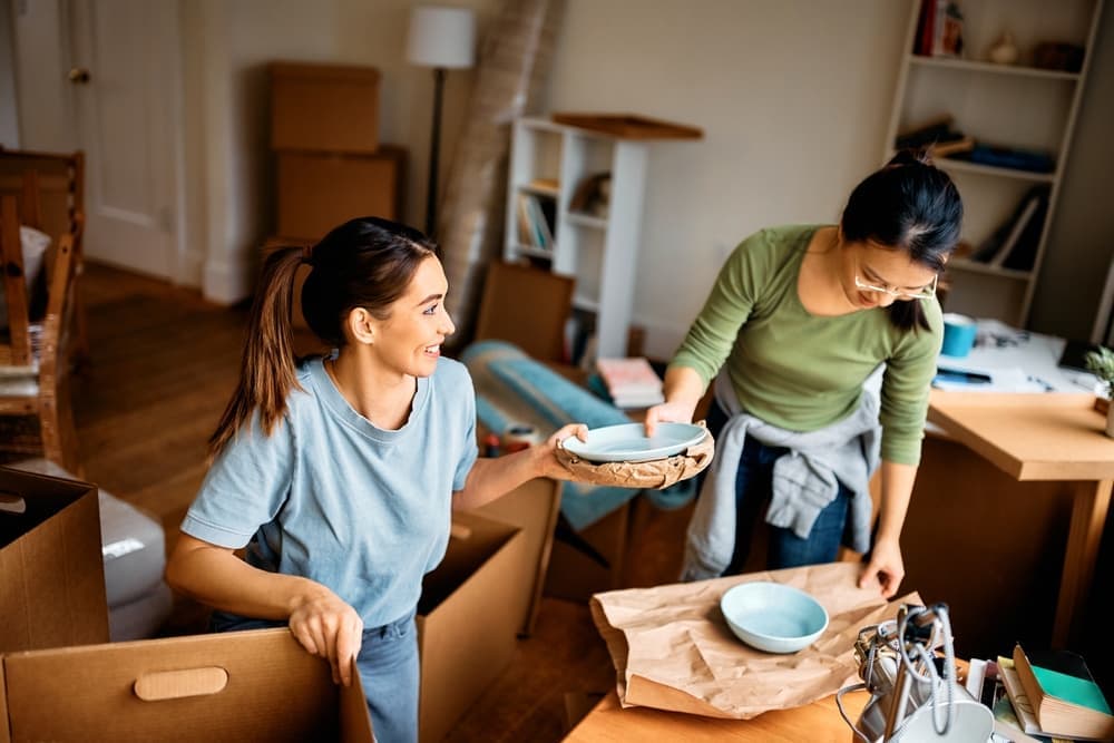 Happy woman and her female friend wrapping fragile things into a paper while preparing to move out of the apartment.