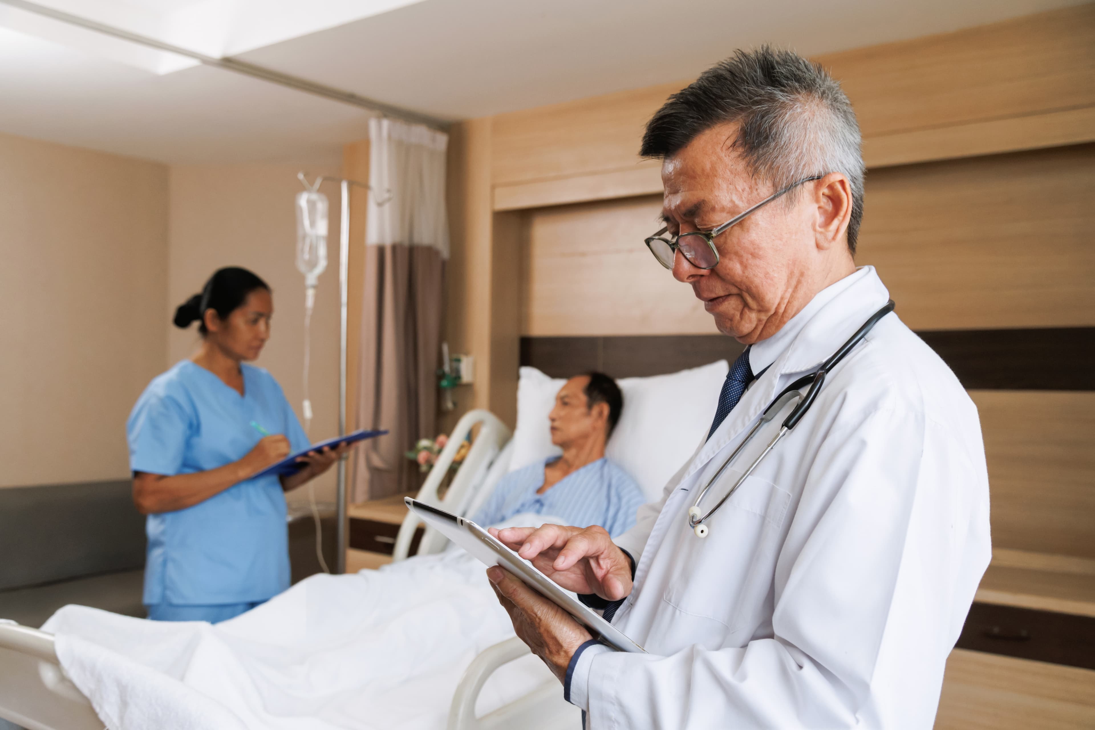Physician reviewing patient records on a tablet at a hospital bedside while a nurse takes notes in the background