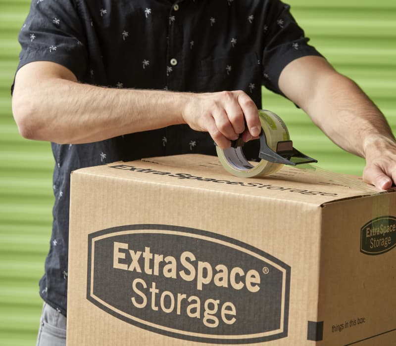 Man packing an Extra Space Storage moving box in front of green storage unit door.