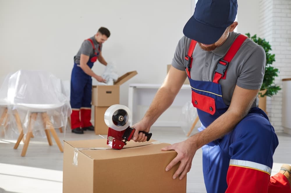 Professional packer taping moving box in foreground with mover in background packing another box for shipping cross-country.