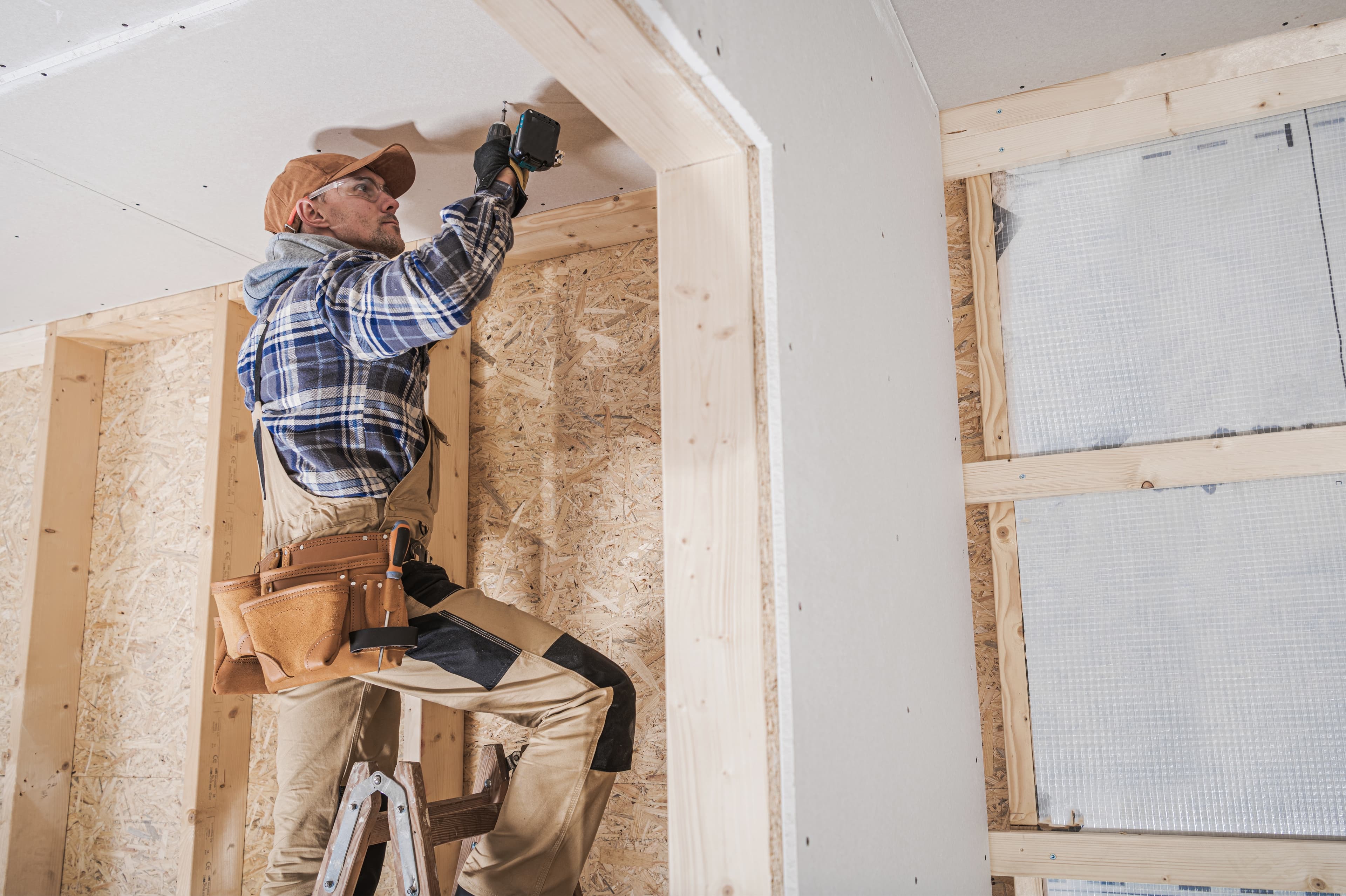 Builder on a ladder using a power drill to install drywall in a wood-framed room under construction