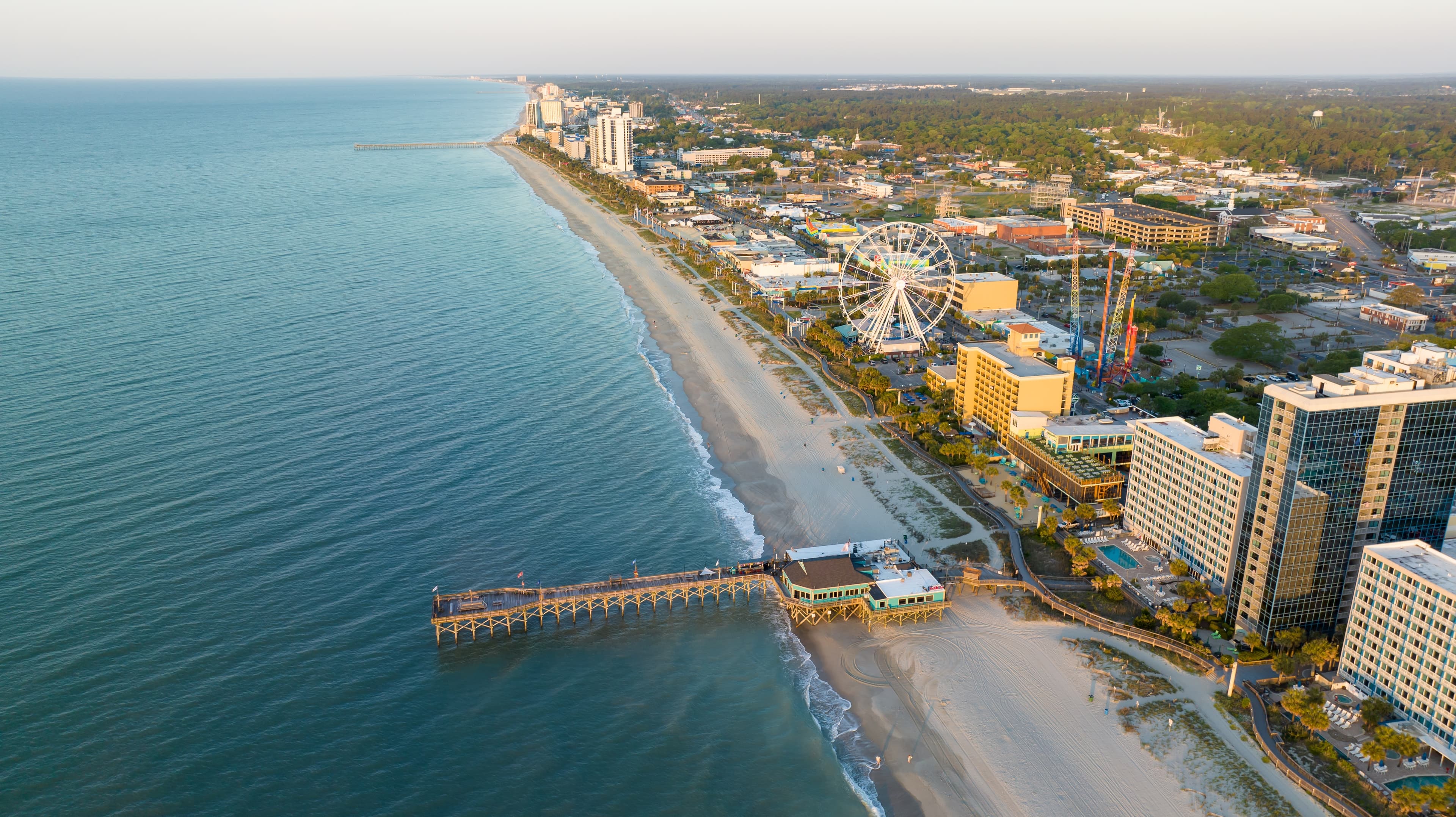 Overhead view of Myrtle Beach, SC featuring skyscrapers, greenspaces, and a beach with a large pier.