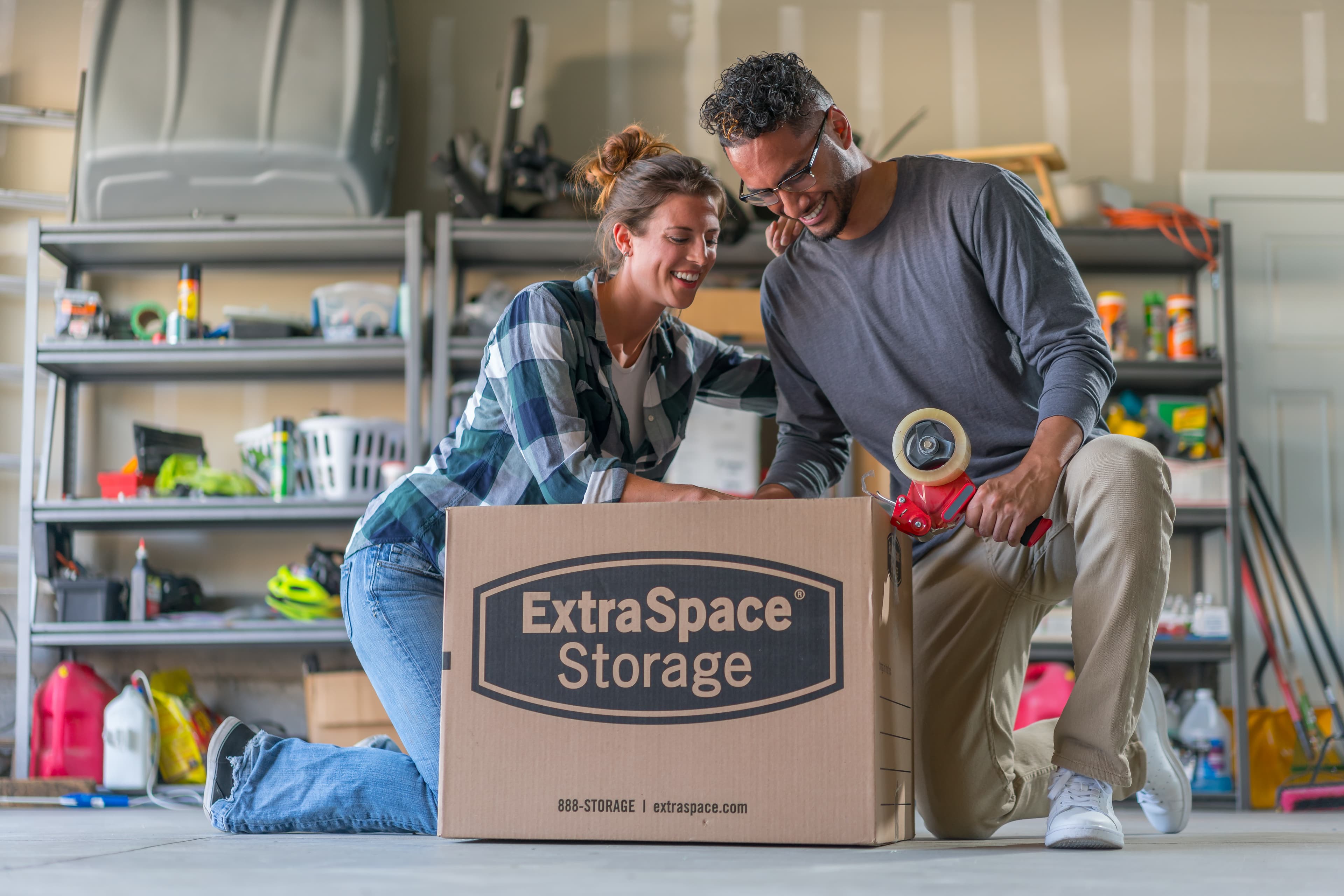 A couple taping a moving box inside of a garage.