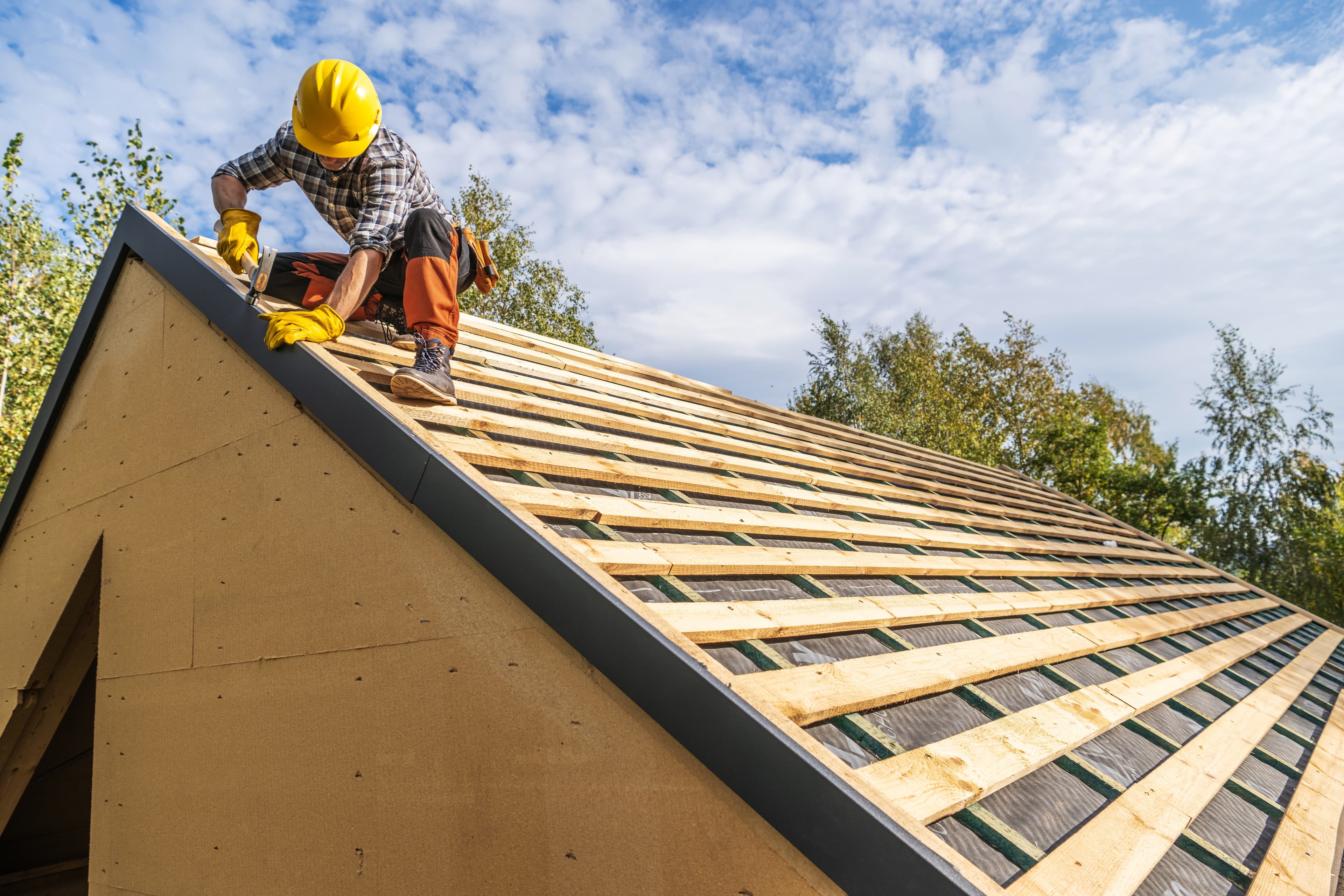Roofer in a hard hat kneeling on exposed wood roof decking, nailing down trim along the ridge