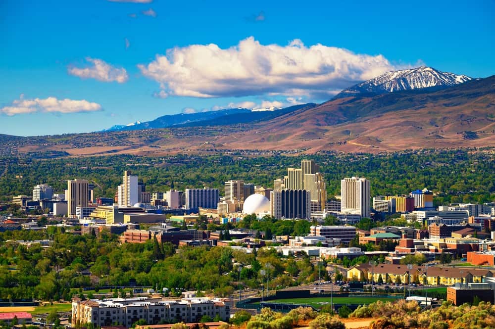 Skyline of Reno, NV featuring the city center with a few tall buildings surrounded by green trees and with mountains and a blue sky with a couple clouds in the background.