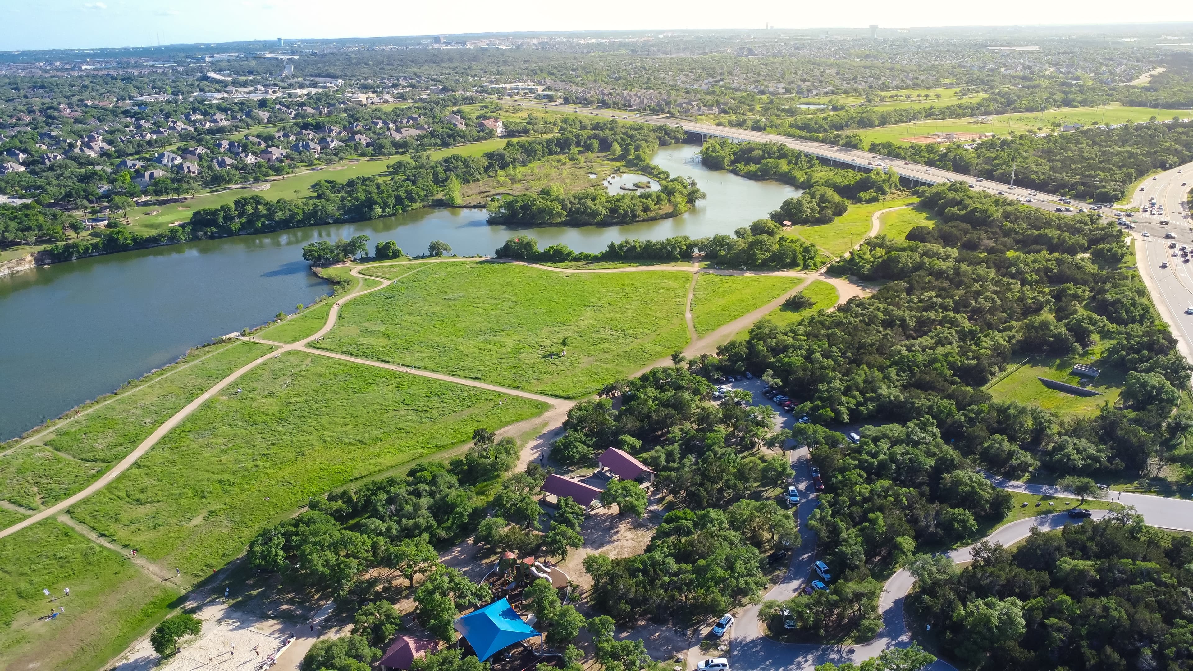 Overhead view of Cedar Park, TX, featuring nice homes, tree-lined streets, fields of grass, and a river on a sunny day.