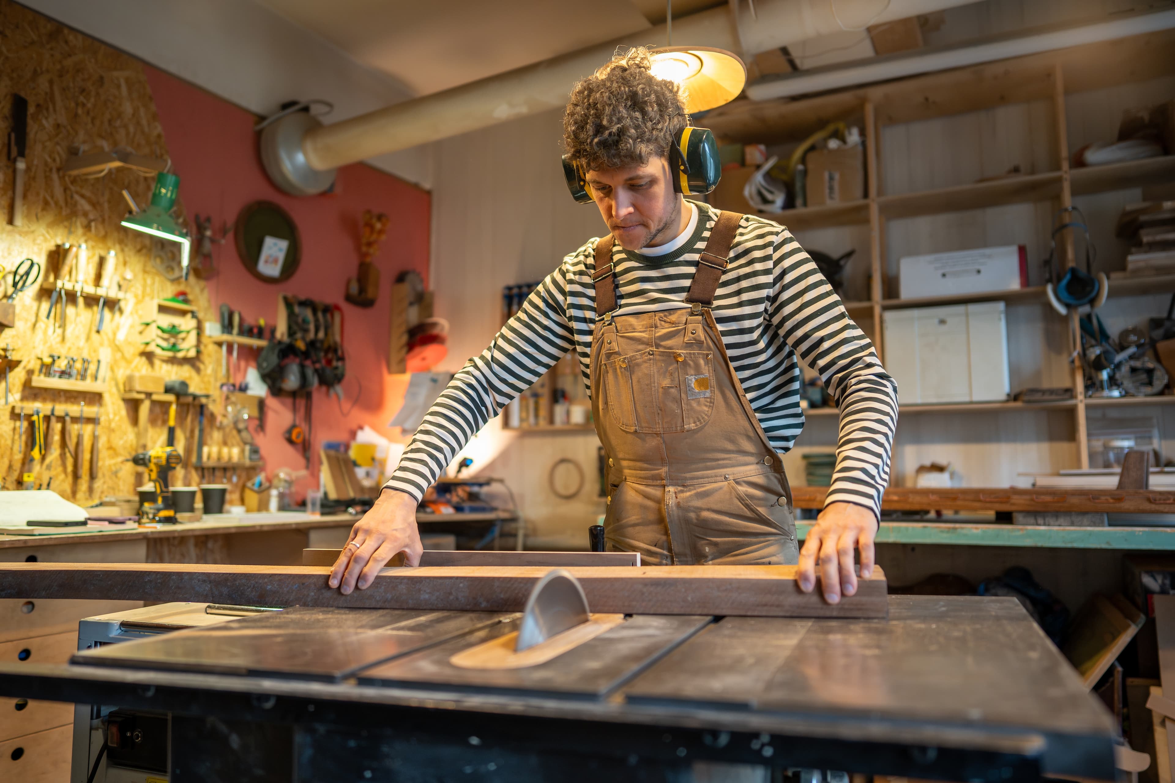 Carpenter wearing ear protection guiding a wood plank through a table saw in a workshop