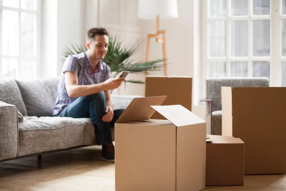 A man surrounded by moving boxes sitting on a couch while looking at his phone