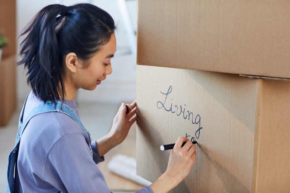 Person with long dark hair in a pony tail and wearing a lavender t-shirt is labeling a box in a stack with a marker