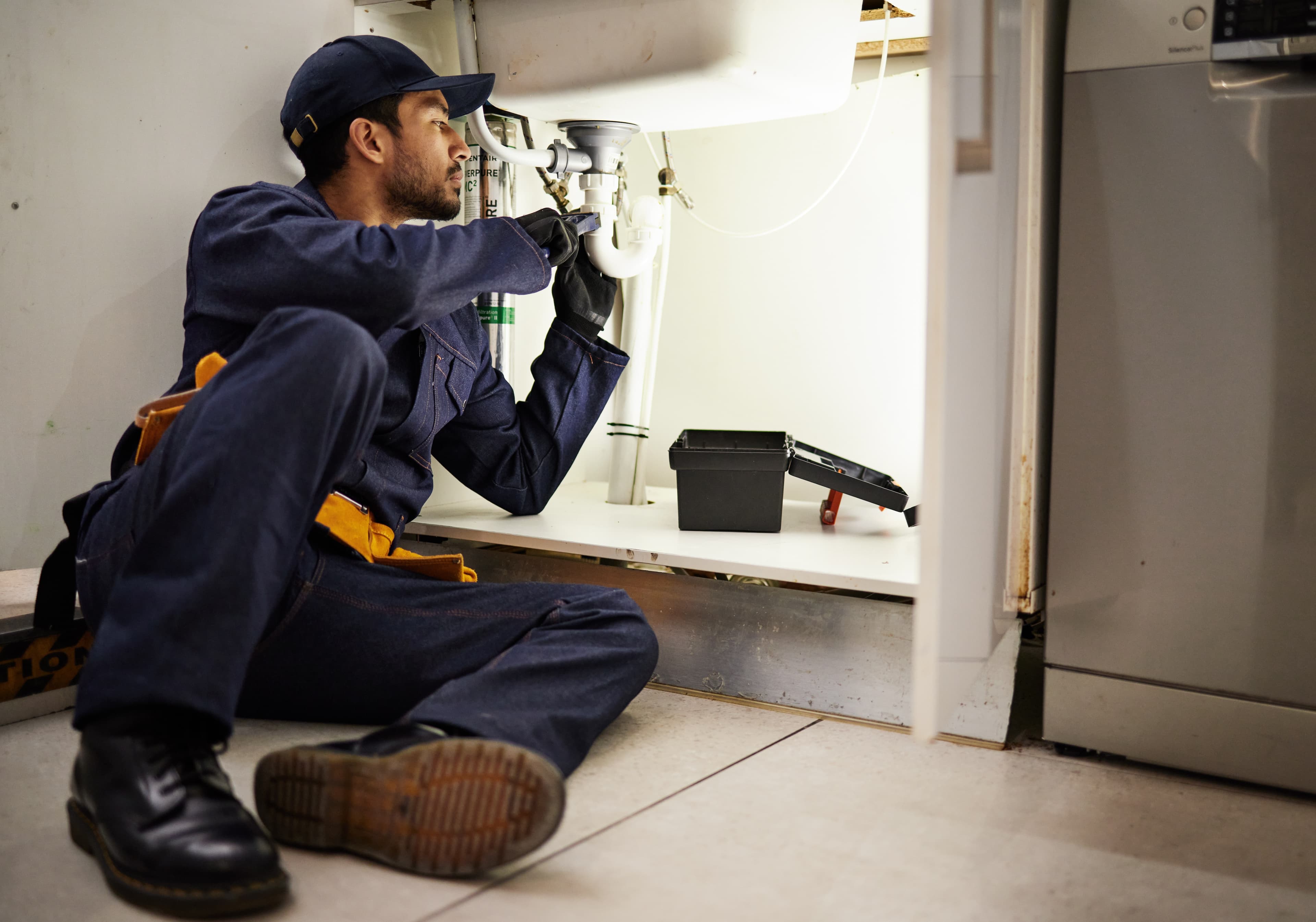 Plumber in coveralls kneeling under a kitchen sink to repair pipes, with a tool kit open on the cabinet floor