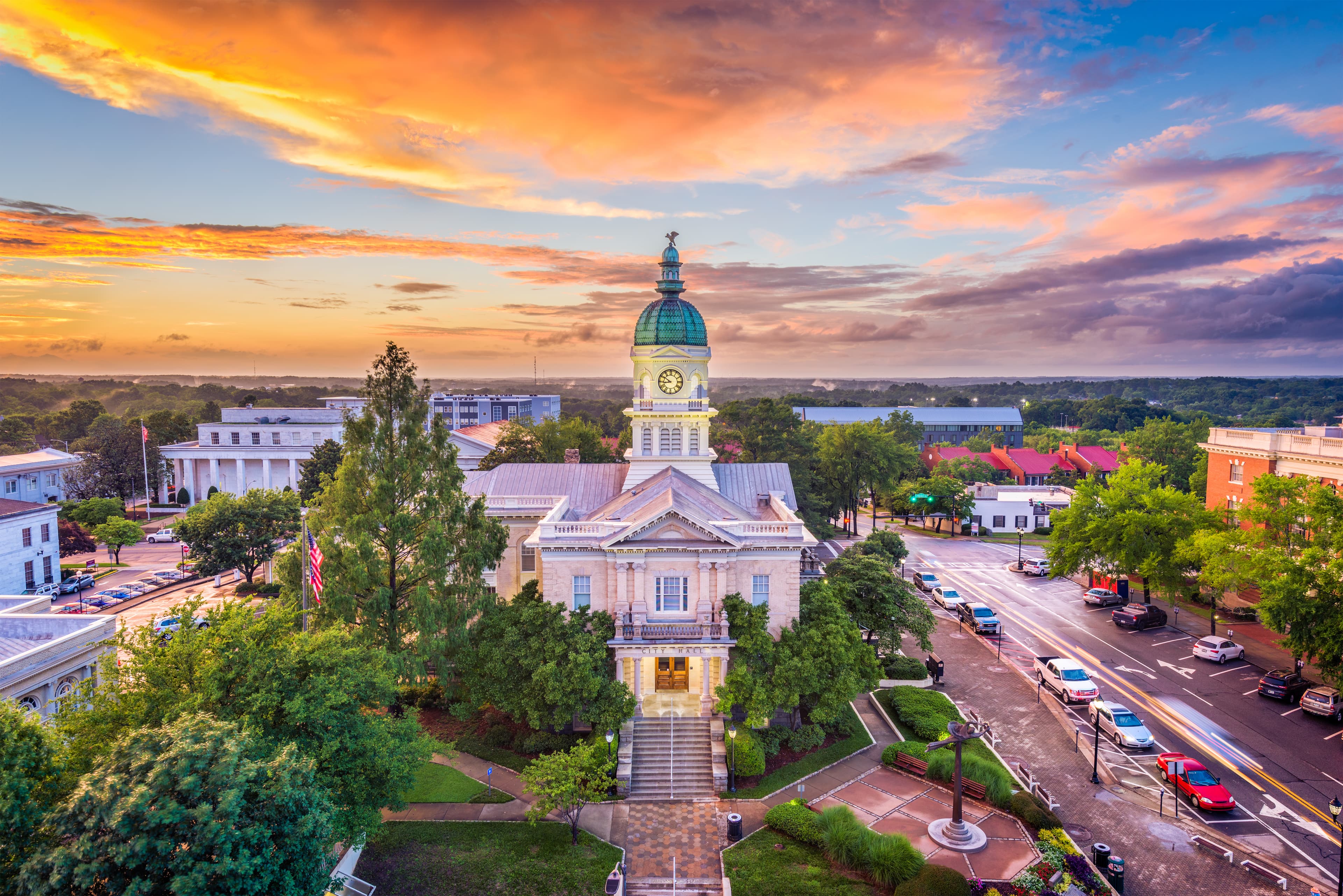 Skyline photo of Athens, GA featuring a busy brick street as well as the city hall surrounded by gardens during sunset.