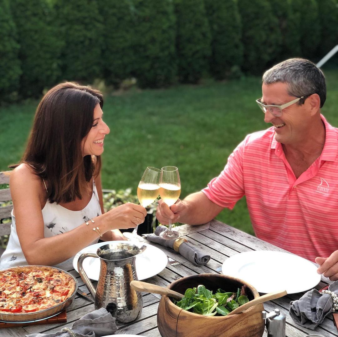 A couple cheer their glasses at a picnic dinner of salad and pizza outside on a wooden table in the sunlight. Photo via Instagram user @caterina.defalco