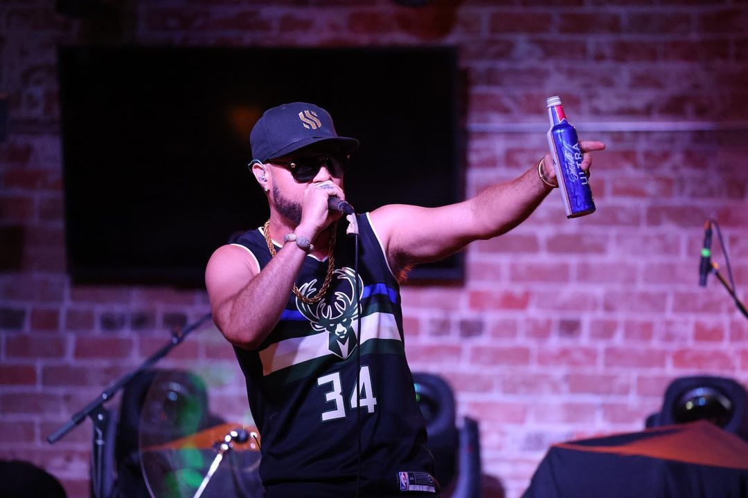 Musician Shy Carter wearing a jersey, baseball hat, and sunglasses, and holding a beverage and microphone in front of a brick wall at an entertainment venue. Photo by Instagram user @shycarterofficial