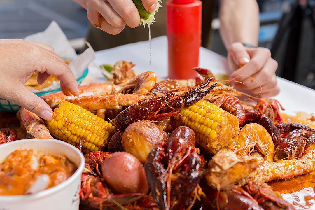 Closeup of crab boil with corn, potatoes and more with two people reaching for it. Photo by Instagram user @boilingcrab
