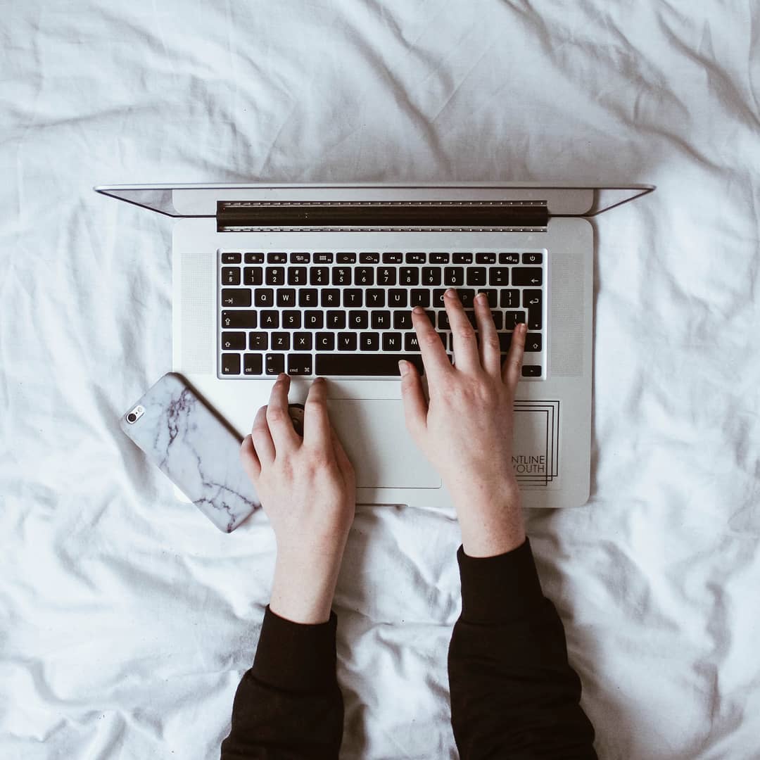 Hands typing on Apple laptop with iPhone nearby. Photo by Instagram user @writersedit