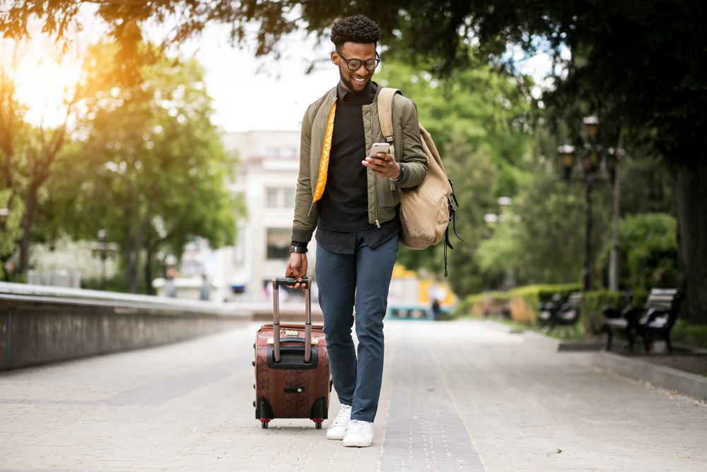 Young man walking with suitcase, looking at phone
