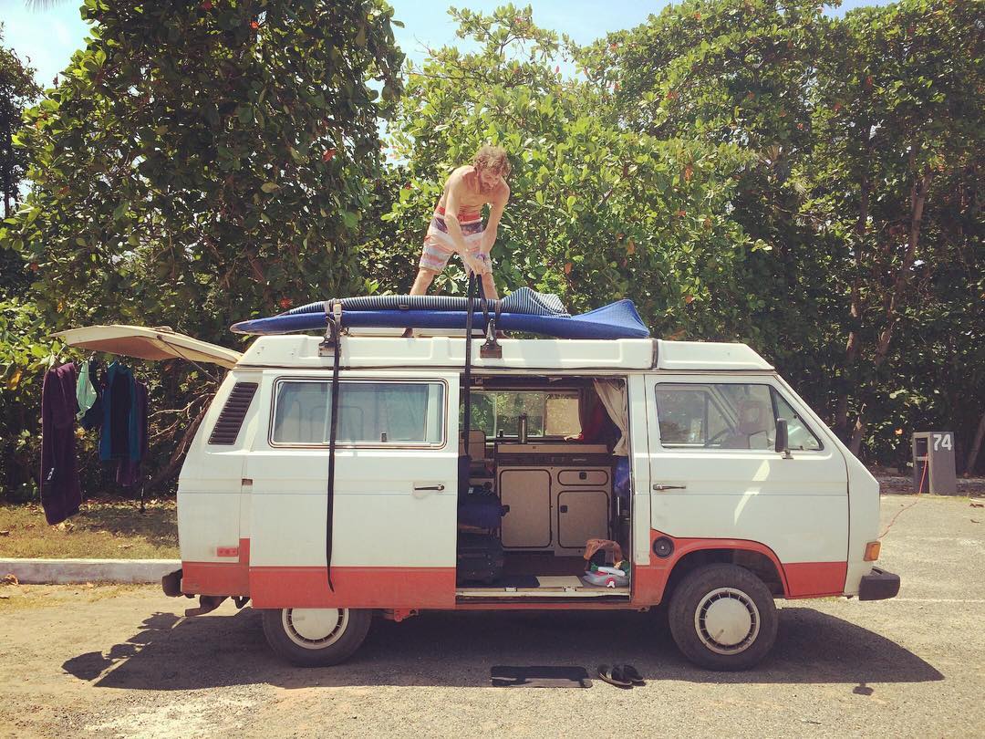 Man strapping surfboard to camper van. Photo by Instagram user @followthesol