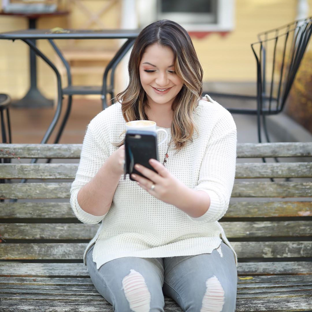 Woman sitting on bench drinking coffee and staring at phone. Photo by Instagram user @allthatjazzhairstylist
