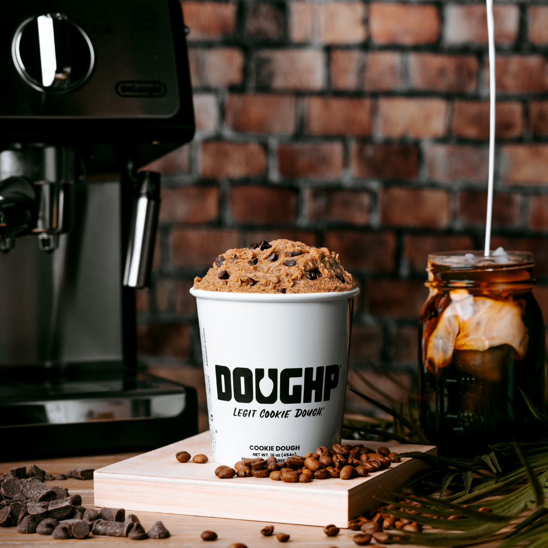 A close up shot of a container DOUGHP edible cookie dough sitting on a countertop next to the coffee machine.