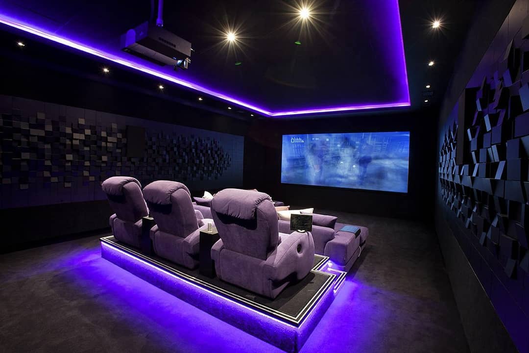 home theater with purple ambient lighting and acoustic pads on walls with large recliners and projector screen photo by Instagram user @allhomeaesthetics
