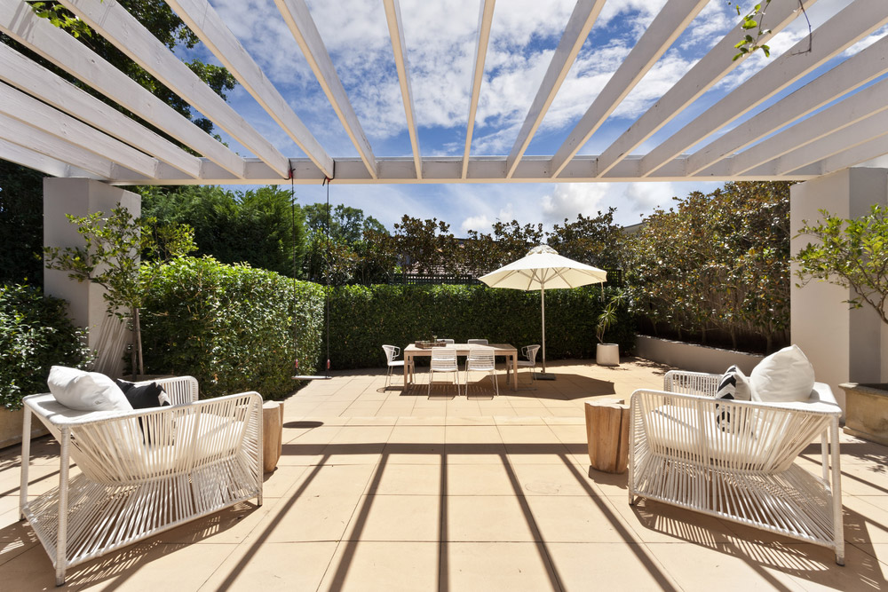 White patio chairs placed underneath a pergola and a dining table with an umbrella in the background