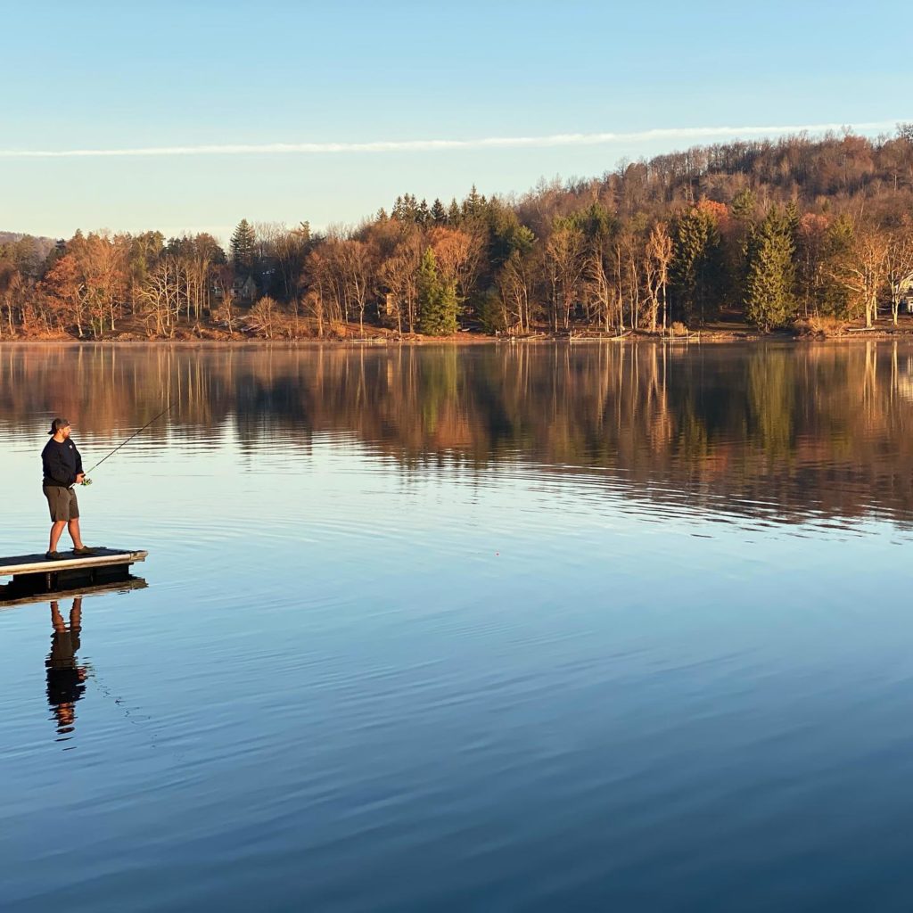 A man fly fishing on a still lake with trees reflecting off the water near the shoreline. Photo via Instagram user @boatsnboardsdeepcreek