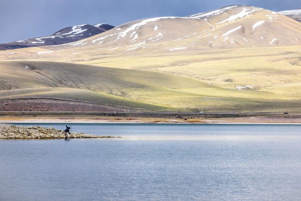 Man fishing from rocky shoreline and large sunlit mountain in background. Photo via Instagram user @jaredssanders