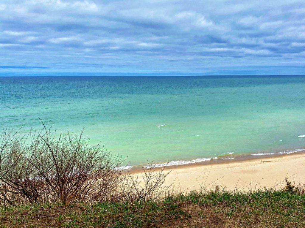 Clear blue green waters with a sandy shoreline and cloudy sky. Photo via Instagram user @ mostly_michigan_