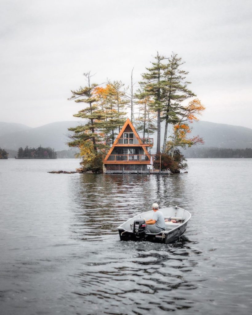 A man boats towards an A-line cabin with evergreens in the middle of a gray, rippling lake on a cloudy day. Photo via Instagram user @will_zimm_