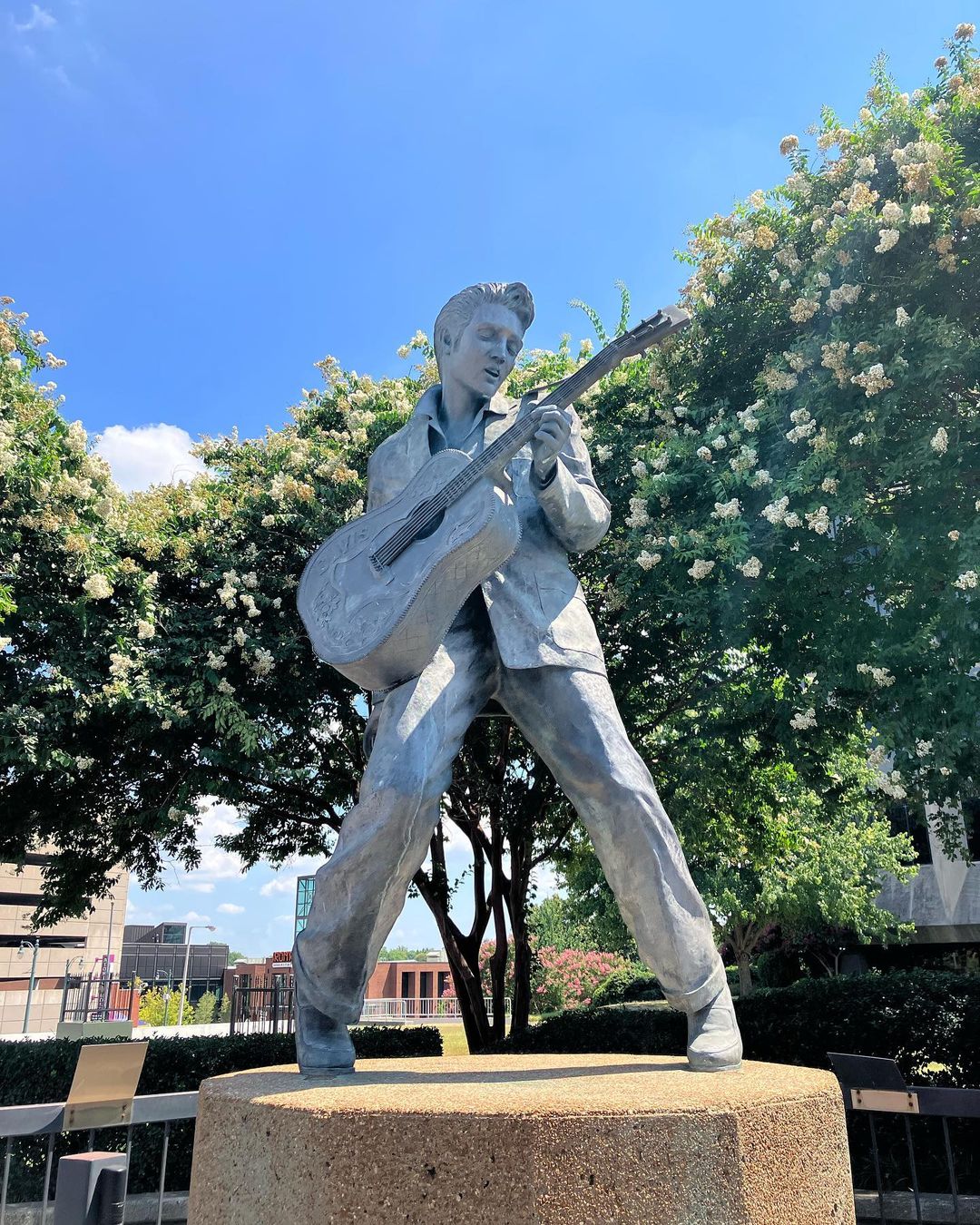 Gray elvis statue posed with a guitar on concrete pedestal surrounded by blooming trees in Memphis. Photo by Instagram user @ladytontour
