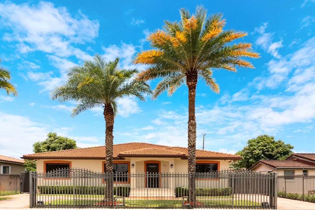 Spanish-style home with two palm trees in front, a fence, and blue skies. Photo by Instagram user @realtorjonny