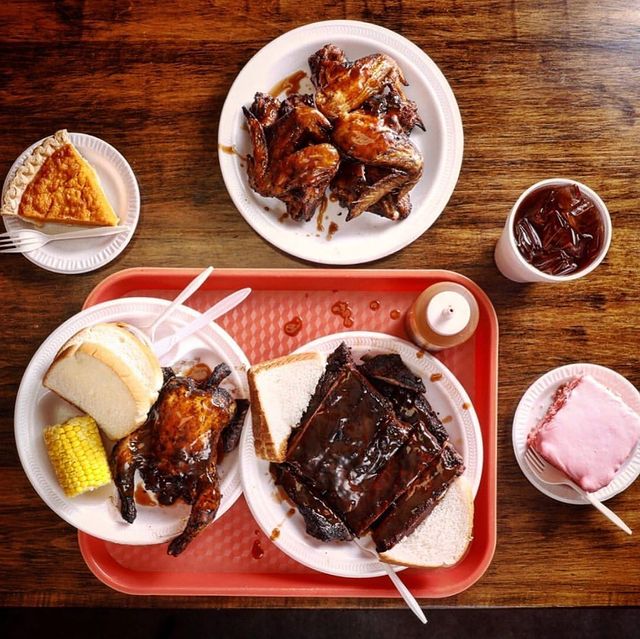 Photo of five paper plates filled with a rack of ribs, wings, and hen smothered in bbq sauce, along with bread, corn, pie, cake, and a soda drink on a red tray from Cozy Corner BBQ restaurant in Memphis. Photo by Instagram user @cozycornerbbq