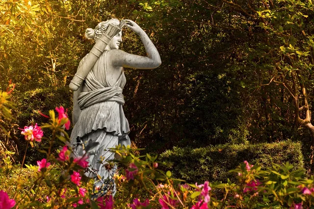 A rock sculpture of a female archer grabbing an arrow from her back sling, surrounded by trees, shrubs, and pink flowers at golden hour. Photo via Instagram user @evrentrx