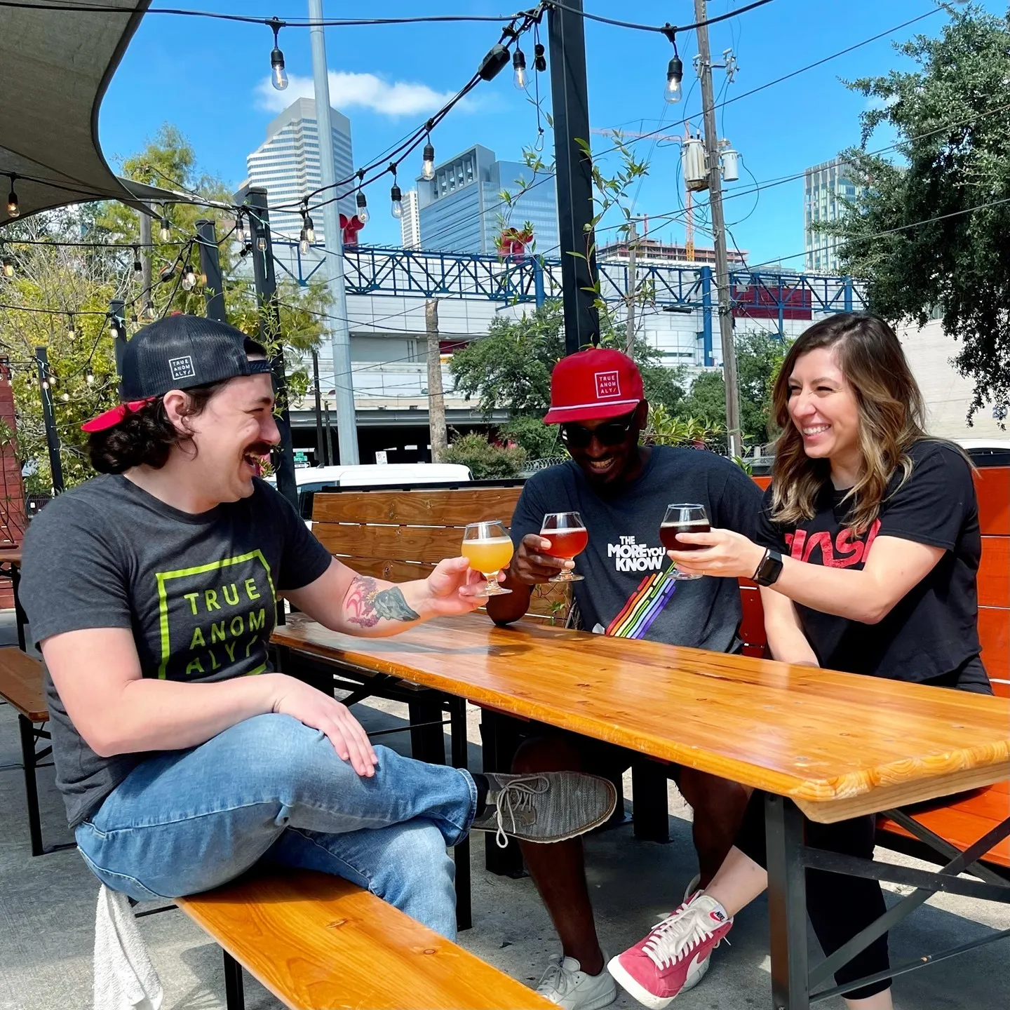 Three young adults cheers their beers at a wooden picnic table underneath string lights with the Houston skyline on a clear sky day in the background. Photo via Instagram user @trueanomalybrewing
