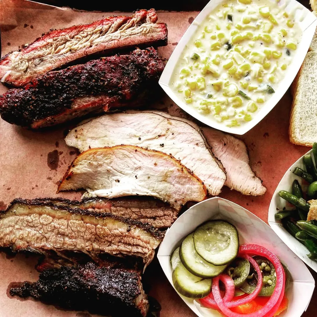 A plate of brisket, ribs, potato salad, pickled onion and cucumbers, and green beans in soft lighting. Photo via Instagram user @willowstexasbbq