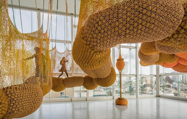 Vistors of the MFA Houston walk through an orange, woven floating walkway made of all fabrics in a bright white room full of windows. Photo via Instagram user @mfahouston