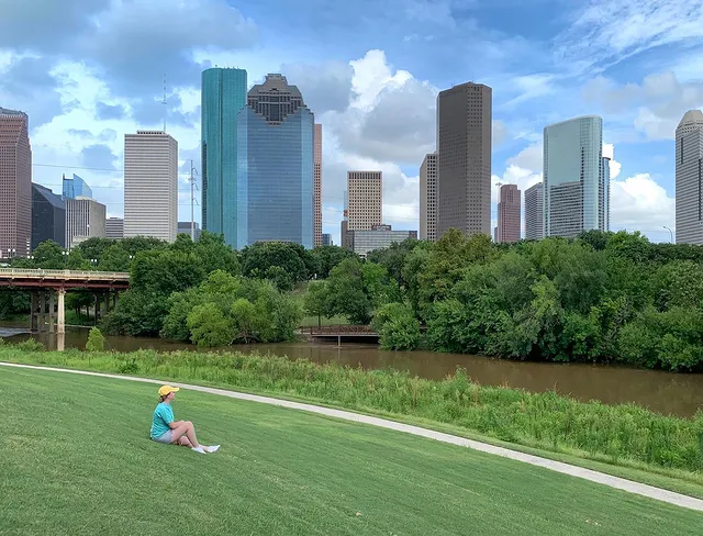 A woman sits on the grass of Buffalo Bayou Park and looks out over the river, trees, and Houston skyline on a partly cloudy day. Photo via Instagram user @myrandaharper