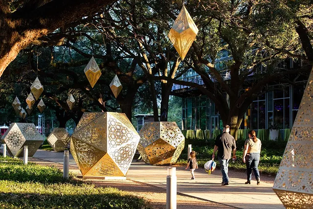 A family of three walks through geometric golden metal sculptures with intricate patterns in the setting sunlight underneath the shade of trees along the sidewalk. Photo via Instagram user @discoverygreen