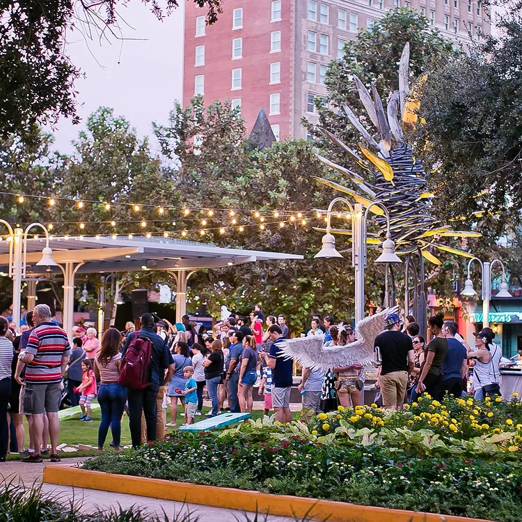 A crowd of people socialize in a downtown Houston sculpture park lit by string lights at dusk with trees, flowers, and buildings around. Photo via Instagram user @downtownhouston